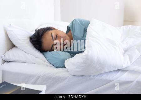 Mid adult Asian woman lying on side wearing light-teal long-sleeve top on bed with white pillow Stock Photo
