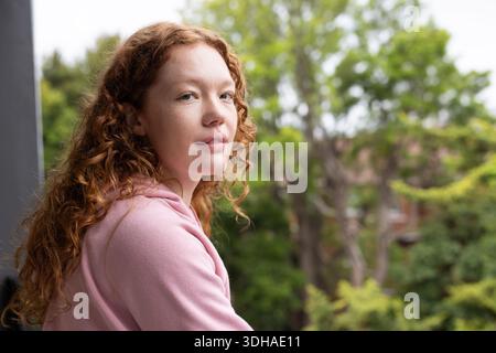 Adult woman leaning on balcony railing near pillar, wearing pink hoodie and gazing at camera Stock Photo
