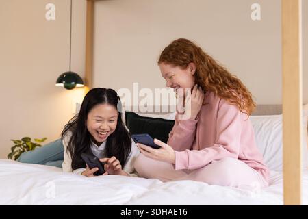 Diverse female friends laughing and sharing phones while lounging on bed with dark green cushion Stock Photo