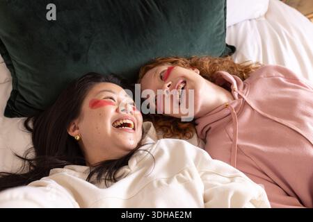 Diverse female friends laughing and lying side-by-side on bed with dark green pillow and face paint Stock Photo