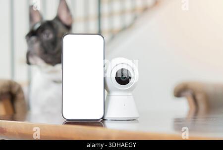 Mini CCTV camera and mobile phone with blank screen setting on wooden table for monitoring of dog at home. Closeup, selective focus. Stock Photo
