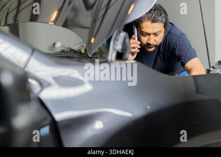 Car repair.  Man working on car while using his mobile phone in garage. At home do it yourself. Examine. Selective focus. Stock Photo