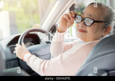 Happy owner. Elder Asian woman with eyeglasses sitting relaxed in her car looking at the camera smiling joyfully. Stock Photo