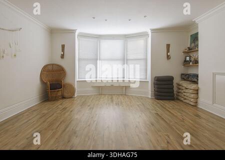 Spacious and bright empty yoga studio featuring hardwood floors, large bay windows, and neatly organized meditation props. Stock Photo