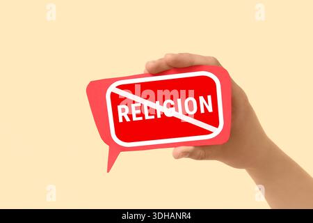Hand holding speech bubble with crossed-out word RELIGION on beige background. Atheism concept Stock Photo