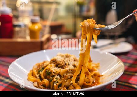 Fork lifting cooked Tagliatelle pasta served with savory meat sauce and cheese in white plate on tartan tablecloth Stock Photo