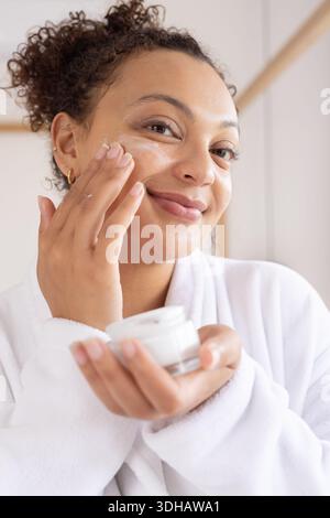 African American woman using cream with tips holding jar wearing white robe gold hoops in bathroom Stock Photo