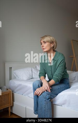 Tired woman sitting on bed in bedroom and blankly looking away, engaged in thinking process. Stock Photo