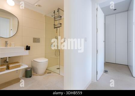 Bright bathroom featuring glass shower, floating vanity, and beige tiles with hallway view Stock Photo