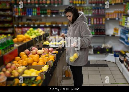 A young woman wearing a gray coat stands at a produce section in a grocery store. She is carefully selecting yellow apples and placing them into a clear plastic bag. Stock Photo