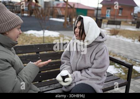 Two friends sit on a bench in a park. They talk while holding cups. The ground is covered with snow. Nearby, there are playground structures and a building in the background. Stock Photo