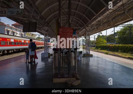 Thrissur, India - March 12, 2023: A group of people, including a man and child, are seen waiting on the platform at Thrissur railway station with a red passenger train stopped alongside, overhead roof structure, signage, and railway tracks visible in daylight. Stock Photo