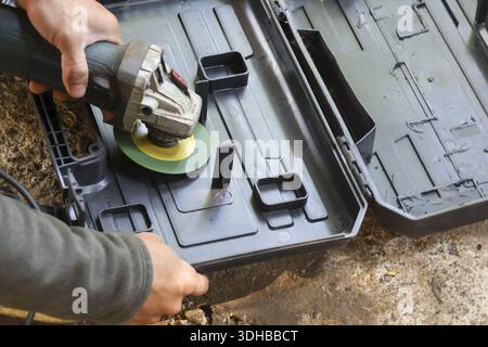 Confident male worker cutting black plastic tool case with electric angle grinder ground carefully, using power equipment for modification task focusi Stock Photo