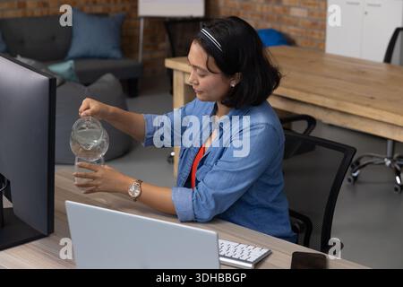 Asian woman pouring water from glass pitcher into tall glass at office desk, wearing denim shirt Stock Photo