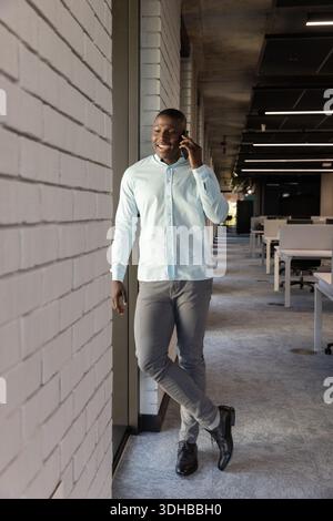 African American man leaning against white brick wall in office holding smartphone in blue shirt Stock Photo