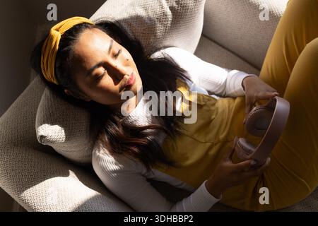 Asian woman reclining on sofa at home wearing mustard overalls holding dusty pink headphones Stock Photo