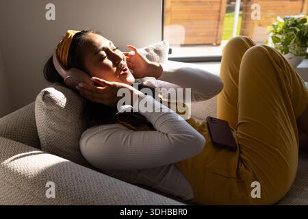 Asian woman lying on sofa at window in mustard overalls holding headphones and smartphone Stock Photo