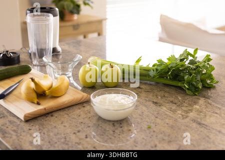 Bananas resting on cutting board with blender, apples, celery and yogurt sitting on granite counter Stock Photo