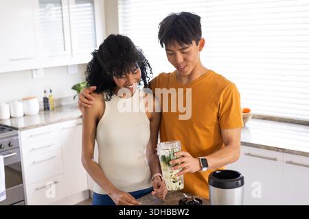 Diverse couple preparing green smoothie in home kitchen, man wearing orange tee holding blender cup Stock Photo