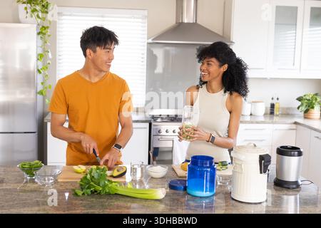 Diverse couple slicing avocados with knife on cutting board, woman holding blender jar in kitchen Stock Photo