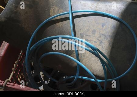 Blue rubber water hose coiled inside dirty old bucket or basin showing neglected messy storage in dim garage atmosphere evoking feeling of abandonment Stock Photo