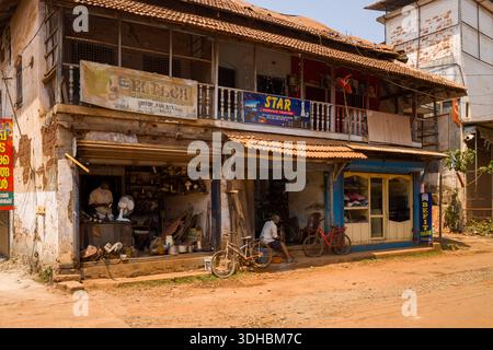 Kannur, India - March 13, 2023: An old two-storey building on a dirt street features an electrical repair shop with a man working inside, a tailor shop with signage, a service centre on the upper floor, bicycles parked outside, and visible shop signs in English and Malayalam under daylight. Stock Photo