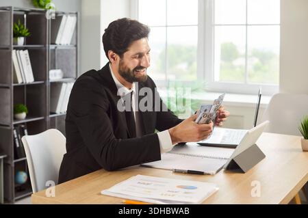 Smiling businessman counting cash by laptop at bright office desk workspace Stock Photo