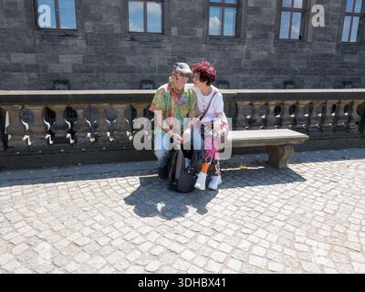 A senior couple sitting on a stone bench next to a historic stone balustrade and a large building wall on a sunny day Stock Photo