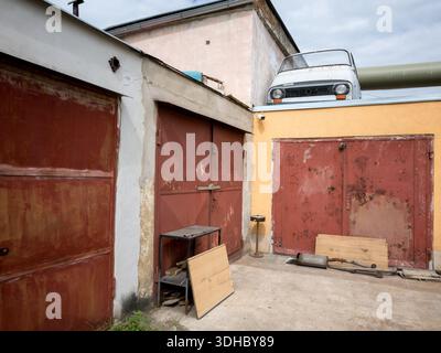A row of old metal garages with rusty red doors, featuring a white Skoda 1203 car front used as a decoration on the roof of a yellow storage unit. Stock Photo