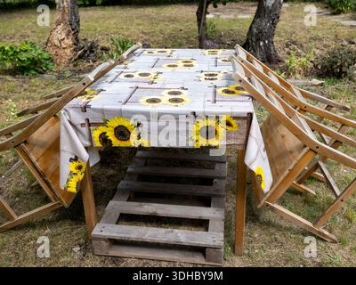 A wooden garden table covered with a sunflower-pattern tablecloth, featuring four chairs tilted against its sides outdoors. Stock Photo