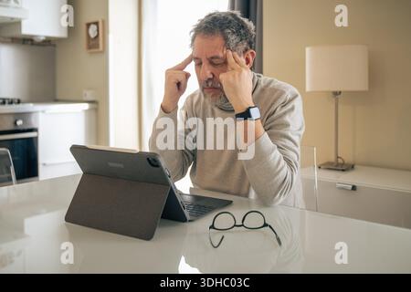 A mature man sitting in a bright kitchen feels a headache while working remotely on a tablet with a keyboard. He is touching his temples with his fing Stock Photo