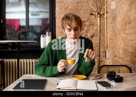 A young man with blonde hair sips coffee in a stylish cafe, focused on his notes. Stock Photo