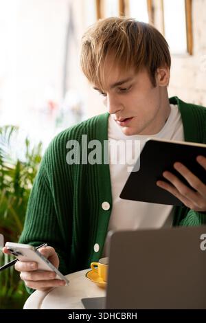 Blonde young man in a green cardigan focuses on his notes while using his smartphone. Stock Photo