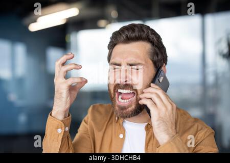 Stressed man shouting loudly with closed eyes and agitated hand gestures while having a difficult conversation on his smartphone in an office setting Stock Photo