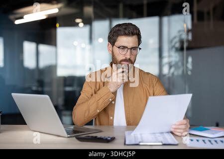 Thoughtful businessman in glasses analyzing financial documents and reports, holding a pen to his chin while working at a desk with a laptop in a modern office, focusing on strategy and planning Stock Photo