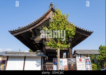 Kyoto, Japan - October 16, 2023: A traditional wooden temple gate with a tiled roof stands behind a tree and a row of colorful posters, with residential buildings and a clear blue sky in the background. Stock Photo