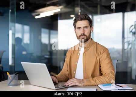 Man with beard and wireless earbuds engaging with his laptop, sitting at a desk in a contemporary office setting, representing focused work and innovative technology use Stock Photo