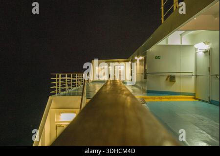 Unknown location - September 13, 2023: The image shows the exterior deck of a ferry at night, featuring railings, illuminated passageways, and safety signage under artificial lighting. Stock Photo