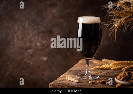 Glass of dark beer with foam head on dark wooden background with empty bottles and beer snacks Stock Photo