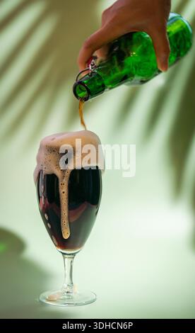 Dark beer pouring from a bottle to a Glass with foam head on colorful green background, bright shadows of palm leaves Stock Photo