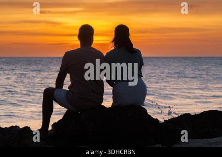 A couple enjoys a serene sunset at Divi Beach, Oranjestad Aruba, with tranquil waves and vibrant sky colors painting the evening. The perfect setting for romance and relaxation. || Model released Stock Photo