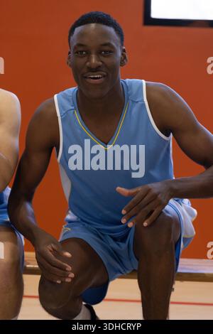 Diverse male teammates resting on wooden bench in gym under scoreboard in light blue uniforms. Athlete, sportsmanship, teamwork, endurance, motivation, training, modern || Model released Stock Photo