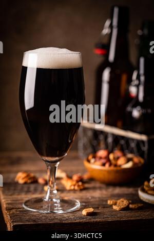Glass of dark beer with foam head on dark wooden background with empty bottles and beer snacks Stock Photo
