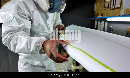 A worker in a protective hazmat suit and respirator carefully applies lime green masking tape to a white foam surfboard blank. The workshop features p Stock Photo
