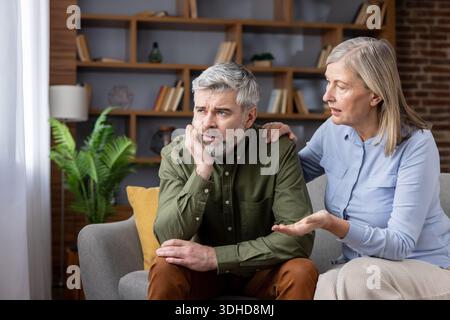 Mature wife comforts distressed husband on the sofa at home, offering empathetic support and attentive listening during a difficult, emotional moment together Stock Photo