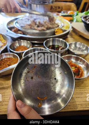 Hand holding an empty Korean barbecue plate with remnants of sauce, surrounded by side dishes and grill Stock Photo
