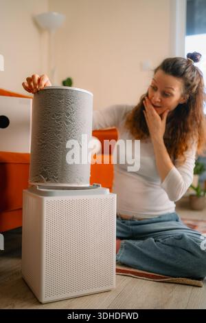 Woman removing a heavily clogged and dusty filter from an air purifier while sitting on the floor, highlighting the importance of regular home appliance cleaning and improving indoor air quality Stock Photo