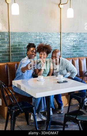 African American friends taking selfie while leaning in and laughing at cafe with coffee cups Stock Photo