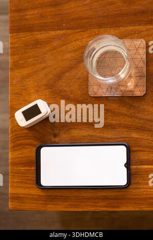 Black smartphone is lying on wood table, showing blank white screen, with glass and oximeter nearby Stock Photo