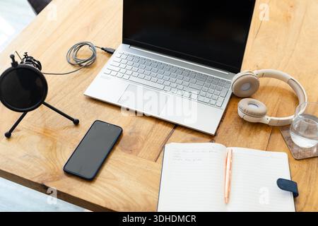 Silver laptop sitting open on wooden desk, catching sunlight, with headphones and mic nearby Stock Photo